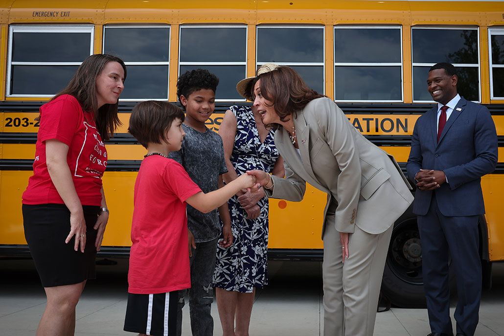 a photo of US EPA Administrator Michael Regan, a white woman with dark hair streaked with grey, and U.S. Vice President Kamala Harris greeting students in front of an electric school bus