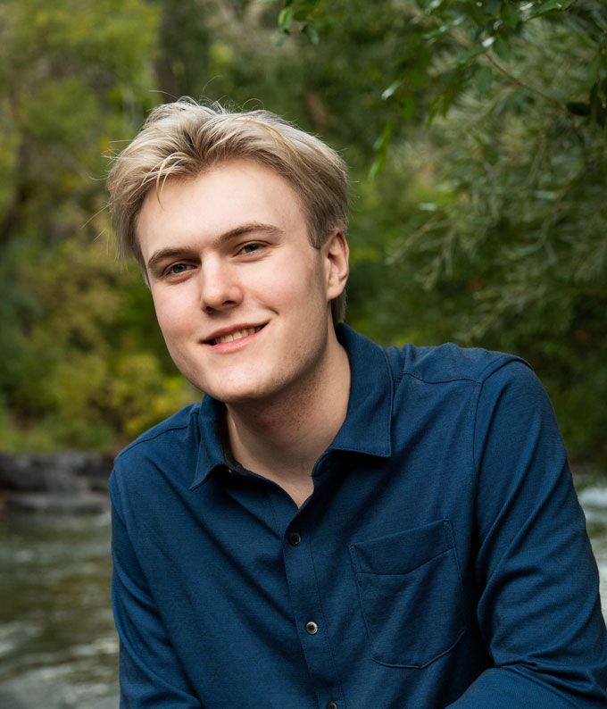 A photo of Liam Sweeney, a young white man with blond hair, smiling at the camera. Behind him are trees and a stream.